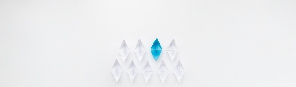 A conceptual image for business leadership featuring a fleet of white origami paper boats following a single bright blue paper boat, symbolizing guidance, strategy, and standing out in the workplace.