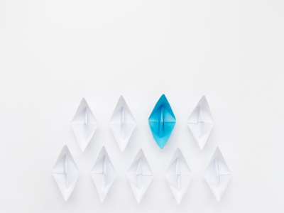 A conceptual image for business leadership featuring a fleet of white origami paper boats following a single bright blue paper boat, symbolizing guidance, strategy, and standing out in the workplace.