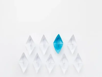 A conceptual image for business leadership featuring a fleet of white origami paper boats following a single bright blue paper boat, symbolizing guidance, strategy, and standing out in the workplace.